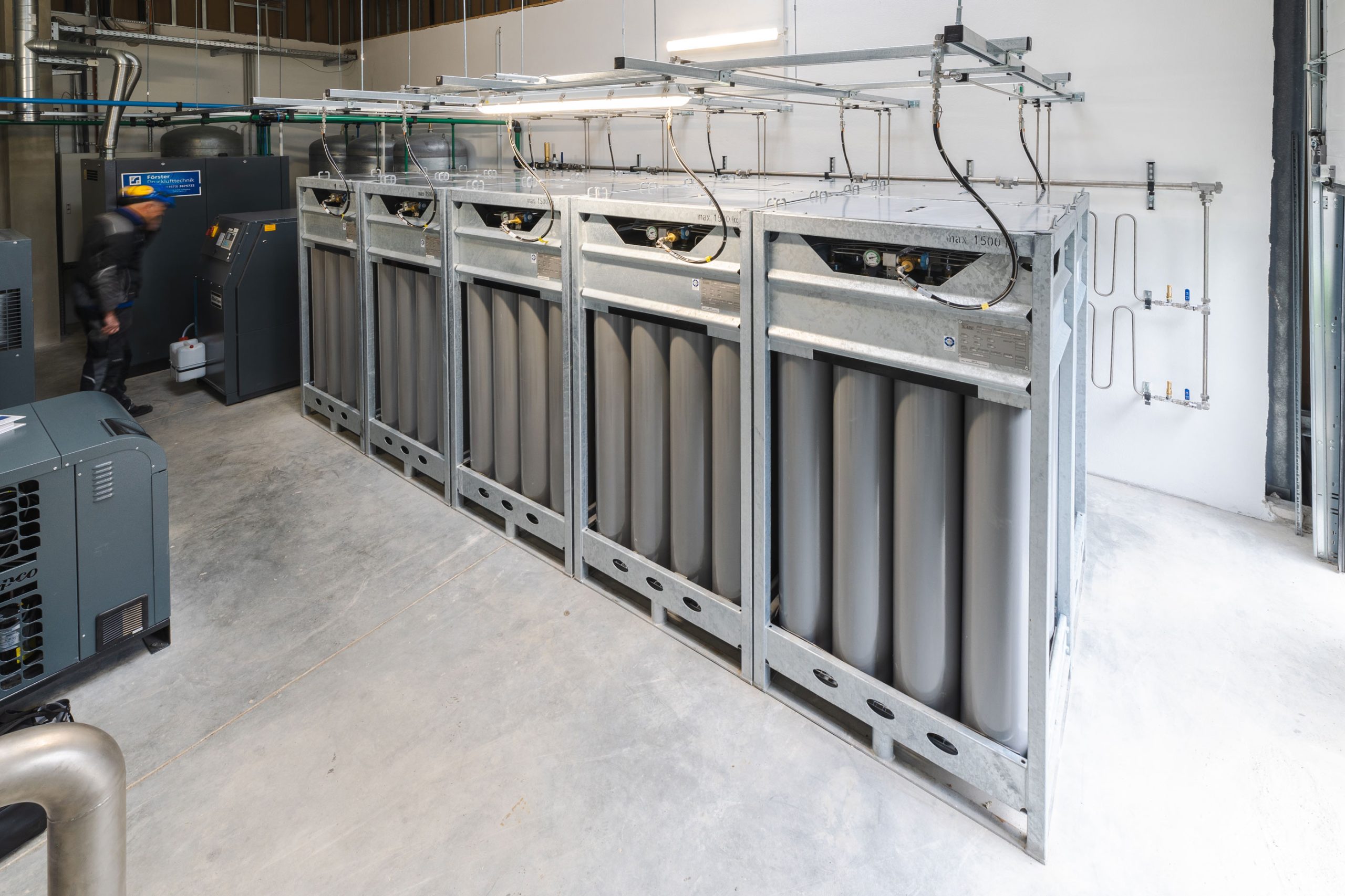 Großer Speicherverbund einer modernen Förster-Druckluft-Anlage in Dresden: mehrere massive Druckbehälter in verzinkten Stahlrahmen stehen in einer sauber strukturierten Industriehalle; rechts und links sind Kompressoren und Leitungen zu sehen, während im Hintergrund ein engagierter Mitarbeiter mit blauem Schutzhelm an der Technik arbeitet.