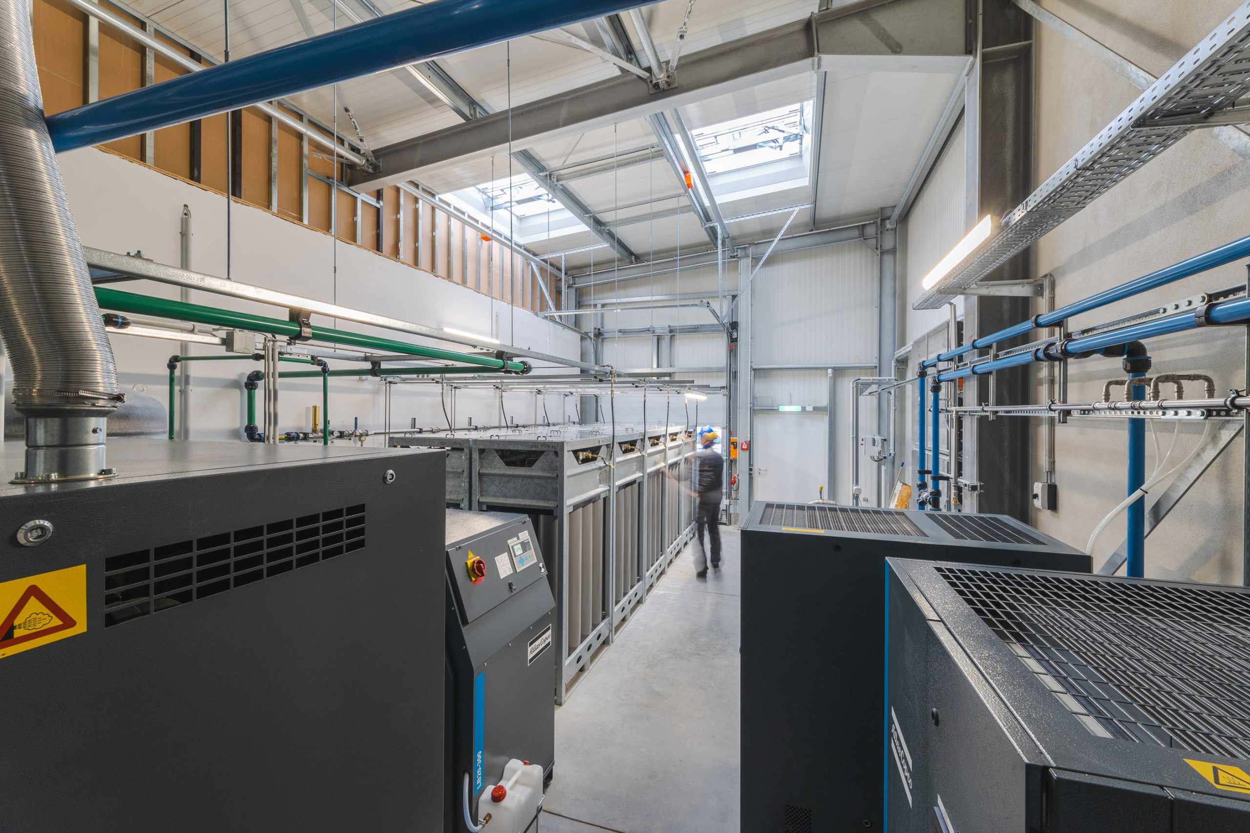 Innenansicht einer modernen Förster-Druckluft-Anlage in Dresden: große Kompressoren, blau-grüne Rohrleitungen und technische Installationen füllen die helle Industriehalle; im Hintergrund arbeitet ein engagierter Mitarbeiter mit blauem Schutzhelm an der Anlage.