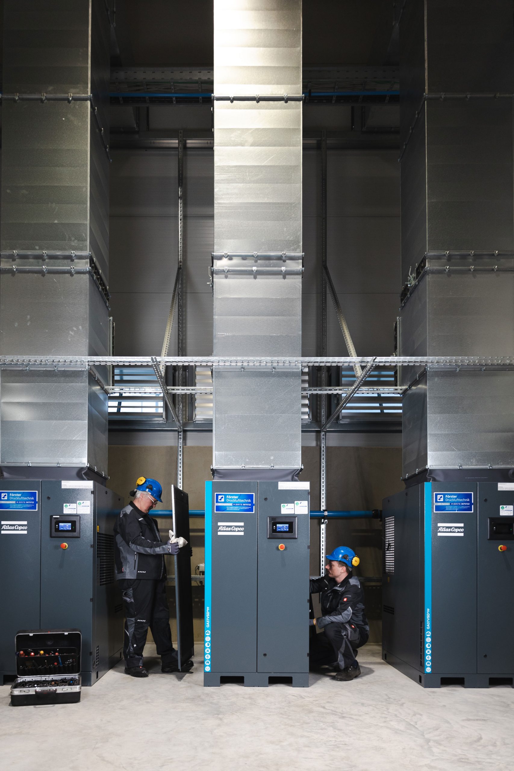Zwei engagierte Förster-Druckluft-Techniker arbeiten in einer modernen Druckluftanlage in Dresden: Links steht ein Mitarbeiter mit blauem Helm und Schutzkleidung vor einem Atlas-Copco-Kompressor und hält eine Geräteabdeckung, während rechts ein weiterer Techniker kniend Wartungsarbeiten durchführt. Aufgereiht stehen mehrere große Kompressoren mit Förster-Druckluft-Beschriftung, darüber verlaufen massive Lüftungs- und Leitungssysteme.
