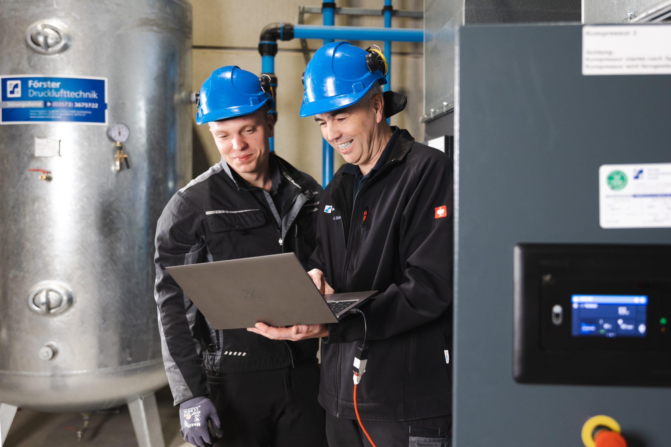 Zwei freundlich lächelnde Förster-Druckluft-Mitarbeiter arbeiten gemeinsam an einer Druckluftanlage in Dresden; sie schauen auf einen Laptop, tragen Förster-Druckluft-Arbeitskleidung und blaue Schutzhelme und sind von modernen Maschinen sowie blauen Leitungen umgeben.