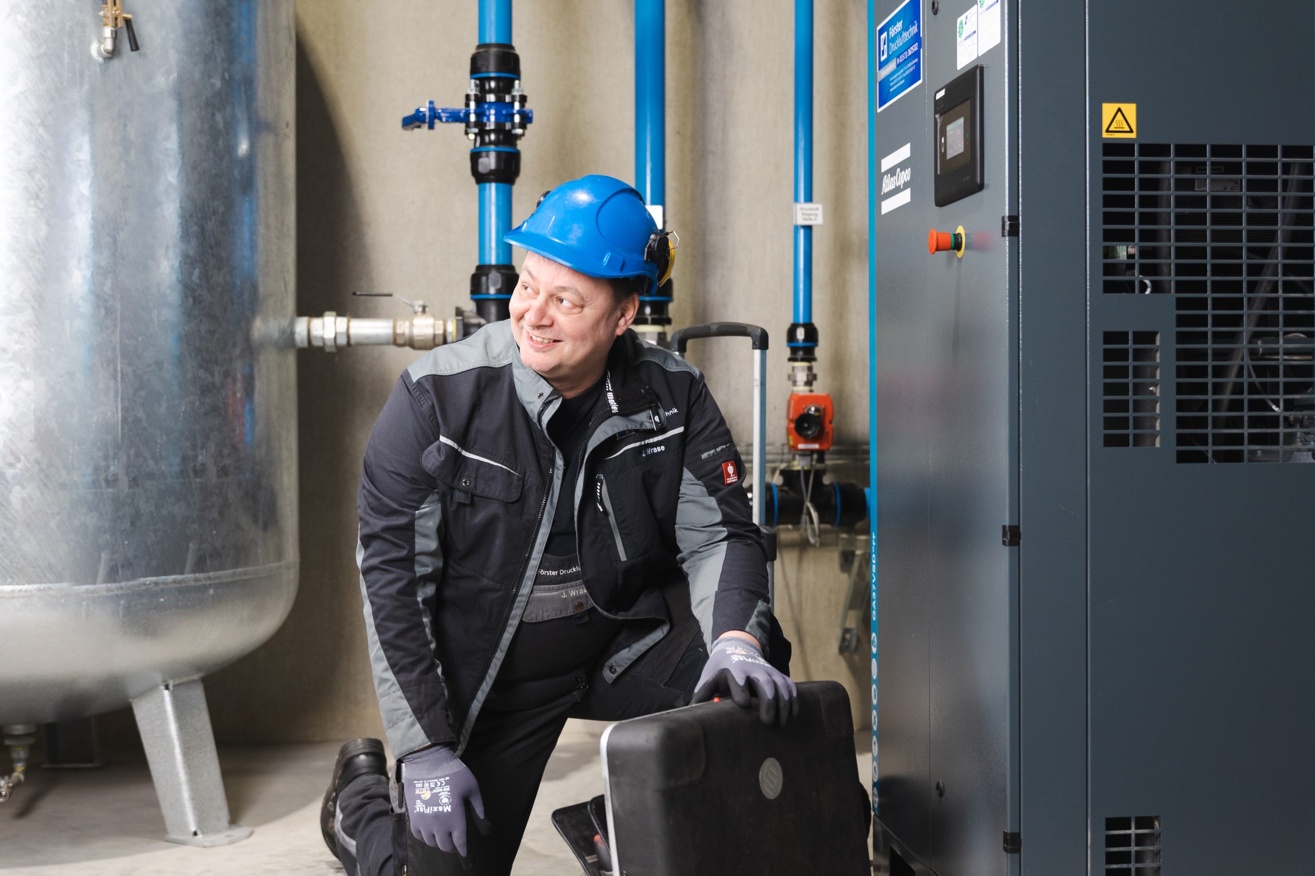 Ein freundlich lächelnder Förster-Druckluft-Mitarbeiter kniet mit seinem Werkzeugkoffer vor einer Druckluftanlage und arbeitet engagiert an der Technik, in einer modernen Förster-Druckluft-Halle in Dresden; blaue Leitungen und ein großer Behälter im Hintergrund.
