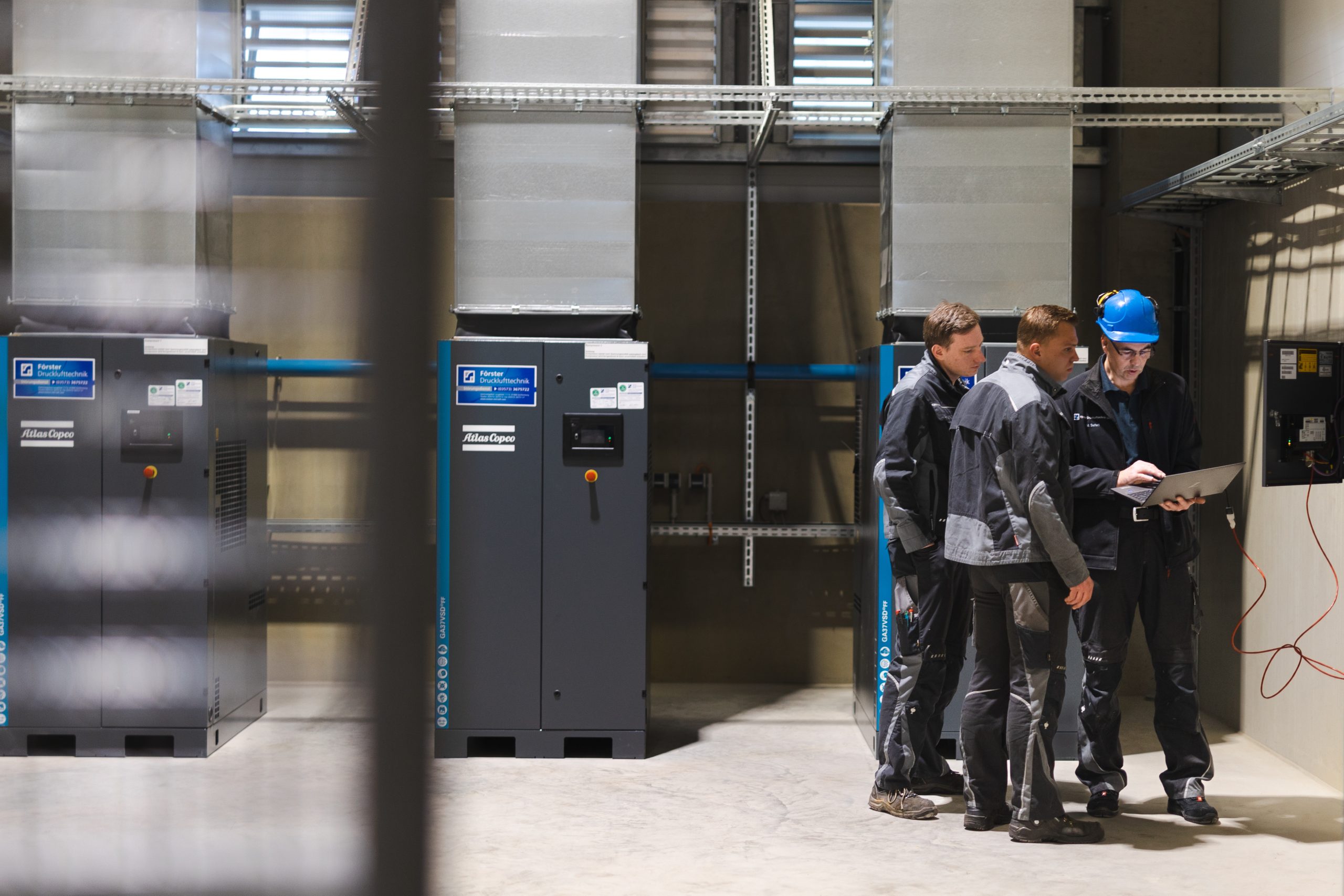 Drei freundliche Förster-Druckluft-Mitarbeiter schauen konzentriert gemeinsam auf ein Laptop und arbeiten in einer modern ausgestatteten Förster-Druckluft-Halle in Dresden an der Anlagenoptimierung.